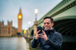 © Itza - Happy young Latin man tourist in casual clothes standing alone against ancient cathedral in London near tower bridge and Big Ben against clock tower and using smartphone
