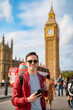 Smiling young Latin male in casual clothes and sunglasses using smartphone while standing on city street during vacation trip to London against Big Ben and clock tower