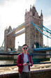 Happy young Latin man wearing sunglasses and red jacket with backpack using smartphone near metal railing of famous Tower Bridge in London