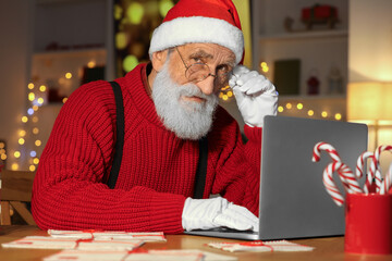 Canvas Print - Santa Claus using laptop at his workplace in room decorated for Christmas