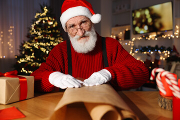 Poster - Santa Claus wrapping gift at his workplace in room decorated for Christmas