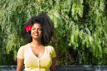 Portrait of young, beautiful, black woman with afro hair, with yellow t-shirt and a red flower in the ear on a background of green plants. Concept beauty, travel, tropical, plants and flowers.