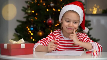 Wall Mural - Portrait of happy girl sits at table with red holiday box and writes letter to Santa Claus on background of Christmas tree. New Year gift