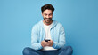 © MP Studio - Young guy smiling holding a smartphone sitting on a blue background