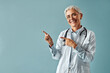 Beautiful confident mature gray-haired woman doctor in white medical coat and glasses standing on light blue background and pointing with fingers to free space in background and smiling.