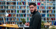© ihorvsn - Male student or researcher in glasses holding laptop computer and typing on keyboard in library room. Handsome Caucasian man turning face to camera, smiling cheerfully in bibliotheca while researching