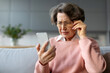 Confused senior woman in eyeglasses squinting eyes reading message on cellphone, having ophtalmic issue problems with vision sitting on couch