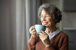 © Home-stock - Happy senior woman drinking coffee looking out window at home, enjoying domestic rest, standing with cup in hands, looking aside and smiling