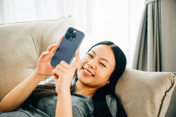 Wall Mural - Young Asian woman relaxes on sofa texting on smartphone. Enjoying relaxation chatting and online shopping. Modern technology for communication and connection.