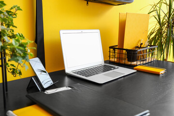 Modern laptop, mobile phone and stationery on dark table near color wall