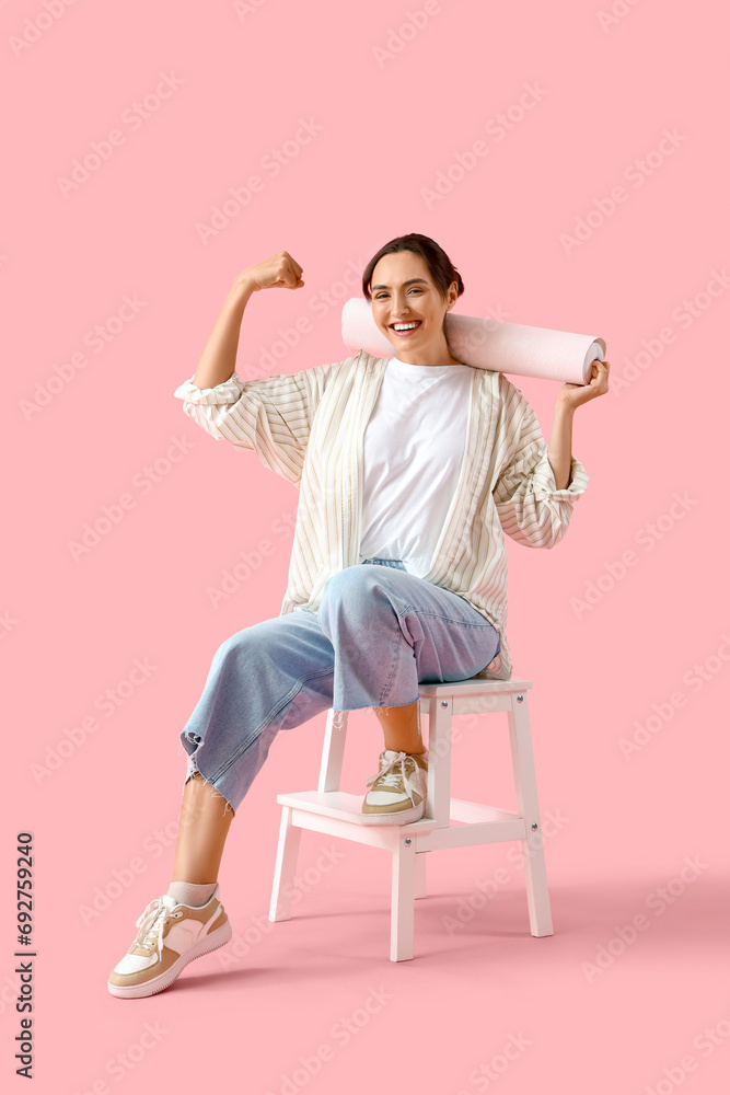 Young woman with wallpaper roll sitting on pink background
