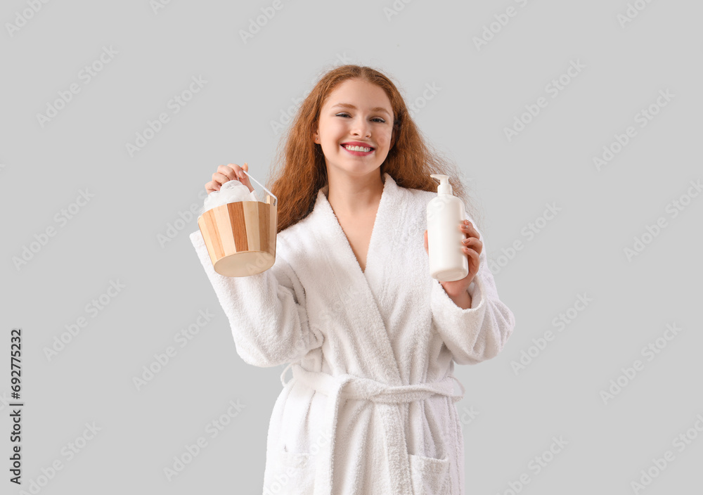 Young woman in bathrobe with shower supplies on light background