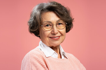 Closeup portrait smiling senior woman, cute happy grandmother wearing sweater, eyeglasses looking at camera isolated on pink background