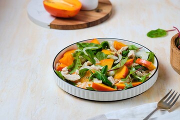 Wall Mural - Green salad with persimmon and boiled chicken in a white plate on a light concrete background. Persimmon recipes.