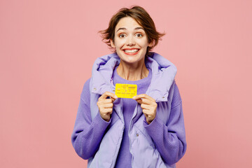 Wall Mural - Young satisfied smiling woman she wear purple vest sweatshirt casual clothes hold in hand mock up of credit bank card isolated on plain pastel light pink background studio portrait. Lifestyle concept.