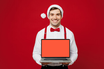 Wall Mural - Merry young fun IT man wear white shirt Santa hat posing hold use work on blank screen laptop pc computer isolated on plain red background studio. Happy New Year Christmas celebration holiday concept.