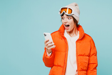 Skier shocked young woman wears warm padded windbreaker jacket hat ski goggles mask hold use mobile cell phone travel rest spend weekend winter season in mountains isolated on plain blue background.