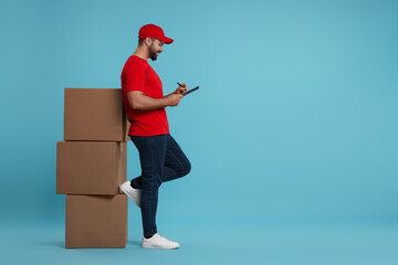 Canvas Print - Happy young courier with clipboard and stack of parcels on light blue background, space for text