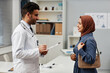 Side view at cheerful Middle Eastern male general practitioner and female patient in brown headscarf meeting in modern clinic office, doctor in glasses holding clipboard with medical card