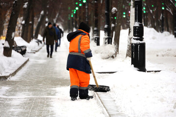 Wall Mural - Communal services worker in uniform with a shovel clears snow on a sidewalk. Woman during snow removal in winter city, street cleaning