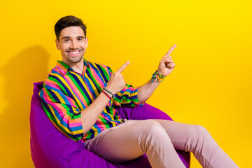 Poster - Photo of young confident young man making promo content point fingers empty space sit pouf courses app isolated on yellow color background