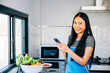 In a cozy kitchen an Asian woman cooks vegetables viewing a cooking tutorial on her smartphone. Smiling and inspecting fruits she merges modern tech with culinary exploration.