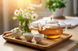 teapot and cups of chamomile tea on wooden tray. Transparent kettle with hot tea on a beautiful background in a room with sunlight from the window
