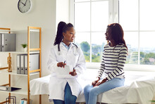 Happy African American Female Doctor And Patient Discussing Treatment. Young Woman Physician In White Medical Coat Sitting On Examination Bed With Young Girl, Talking About Her Therapy, Giving Advice