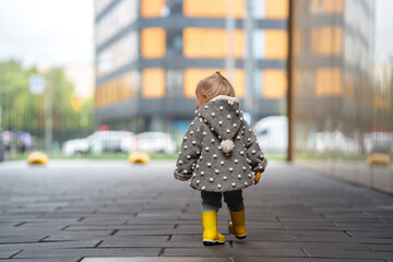 Wall Mural - A cute stylish two-year-old toddler girl kid in a gray coat and yellow boots walks outdoors in the city in autumn season 