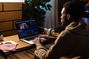 Wall Mural - Attractive young African American filmmaker, using his phone to storyboard ideas and communicate with crew members while editing footage on his laptop.