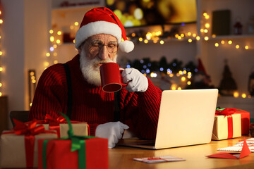 Canvas Print - Santa Claus using laptop and drinking hot beverage at his workplace in room decorated for Christmas