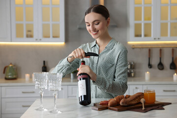 Poster - Romantic dinner. Happy woman opening wine bottle with corkscrew at table in kitchen