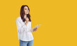 © Studio Romantic - Pretty brunette woman in white shirt and jeans standing isolated on yellow copy space studio background, holding pen and clipboard, thinking what to write and looking away with pensive face expression