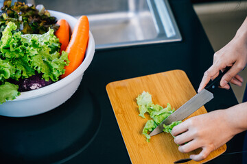 Wall Mural - Close-up of an unrecognizable woman in a blue apron cutting fresh vegetables for a healthy salad. Illustrating the housewife's preparation of a nutritious meal in a modern kitchen.