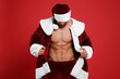 Attractive young man with muscular body in Santa costume on red background
