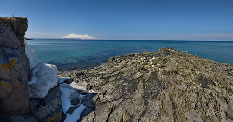 Wall Mural - Russia. The Far East, the Kuril Islands. View of the cold expanse of the Sea of Okhotsk against the background of snow-capped volcanoes surrounded by basalt rocks.