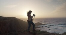 Epic And Cinematic Shot Of Female Adventure Or Travel Photographer Stand On Edge Of Cliff In Evening Sunset Light. Wind Blow Her Hair Around. Explore Exotic Travel Destination And Create Content