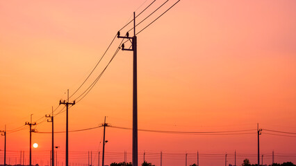 Silhouette row of electric power poles with cable lines against orange sunset sky background 