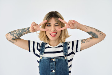 Funny gen z blonde young woman, girl with short blond hair tattoos wearing striped t-shirt and denim dress having fun showing tongue and peace signs over face isolated on white background. Portrait