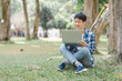 © M+Isolation+Photo - Young Asian student is sitting on the grass, focused on his laptop screen, probably working on his homework or studying.