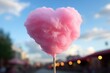 A fluffy pink cotton candy in the shape of a heart on a stick with a blurred street scene in the background.