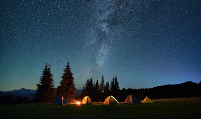 night camping in mountains under starry sky. tourist tents in campsite near burning campfire under b