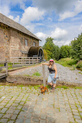 Wall Mural - Smiling mature woman with her brown dachshund standing in front of old Eper or Wingbergermolen water mill in background, sunny day blue sky with clouds in Terpoorten, Epen, South Limburg, Netherlands