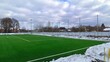 The stadium is located among trees, buildings and is surrounded by a metal fence with high netting at the ends. The soccer field is covered with artificial grass with markings. In winter the snow