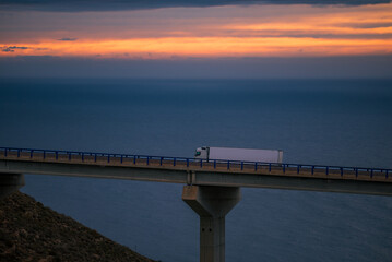 Poster - Truck with refrigerated trailer driving over a bridge with the sea and a dramatic sunrise sky on the horizon.