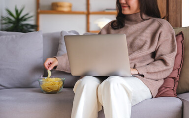 Wall Mural - Closeup image of a young woman eating potato chips while working or watching movie on laptop at home