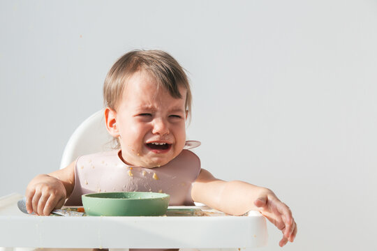 cute crying baby sitting eating lunch in high chair at home on white background, hysterics and tears in a 1 year and 3 month old child, close up portrait.