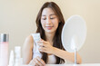 © KMPZZZ - Healthy skin care, smile asian young woman, girl read label to use cream on the tube before applying moisturizer, putting cream treatment on face, makeup routine in the morning at home. Facial Beauty.