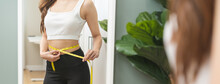 Weight Lose, Loss Concept. Slim Body Asian Young Woman Hand Use Tape Measuring Around Waistline In Fit Sports. Girl Looking Reflect In Mirror At Home. Healthy Nutrition, Fitness For Wellbeing Beauty.
