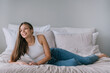 © Iona - Relaxed young woman lounging on a bed, casually dressed in a white tank top and blue jeans, with a serene smile, embodying comfort and effortless style in a cozy setting.